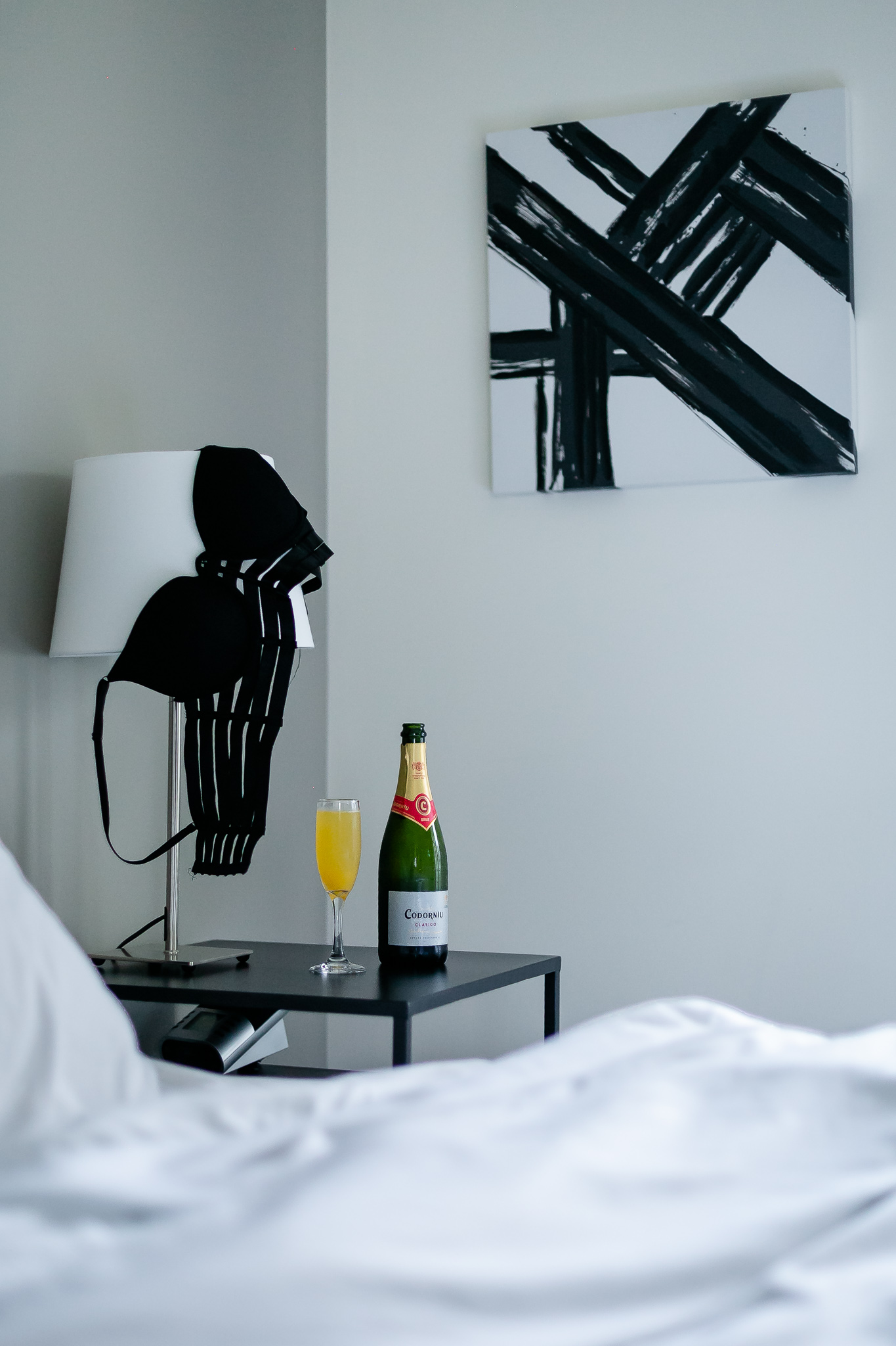 Image is taken in a bedroom. There is a nightstand in the corner with a champagne bottle and a glass with orange juice. On the nightstand is also a lamp, with a black bra tossed over it. 